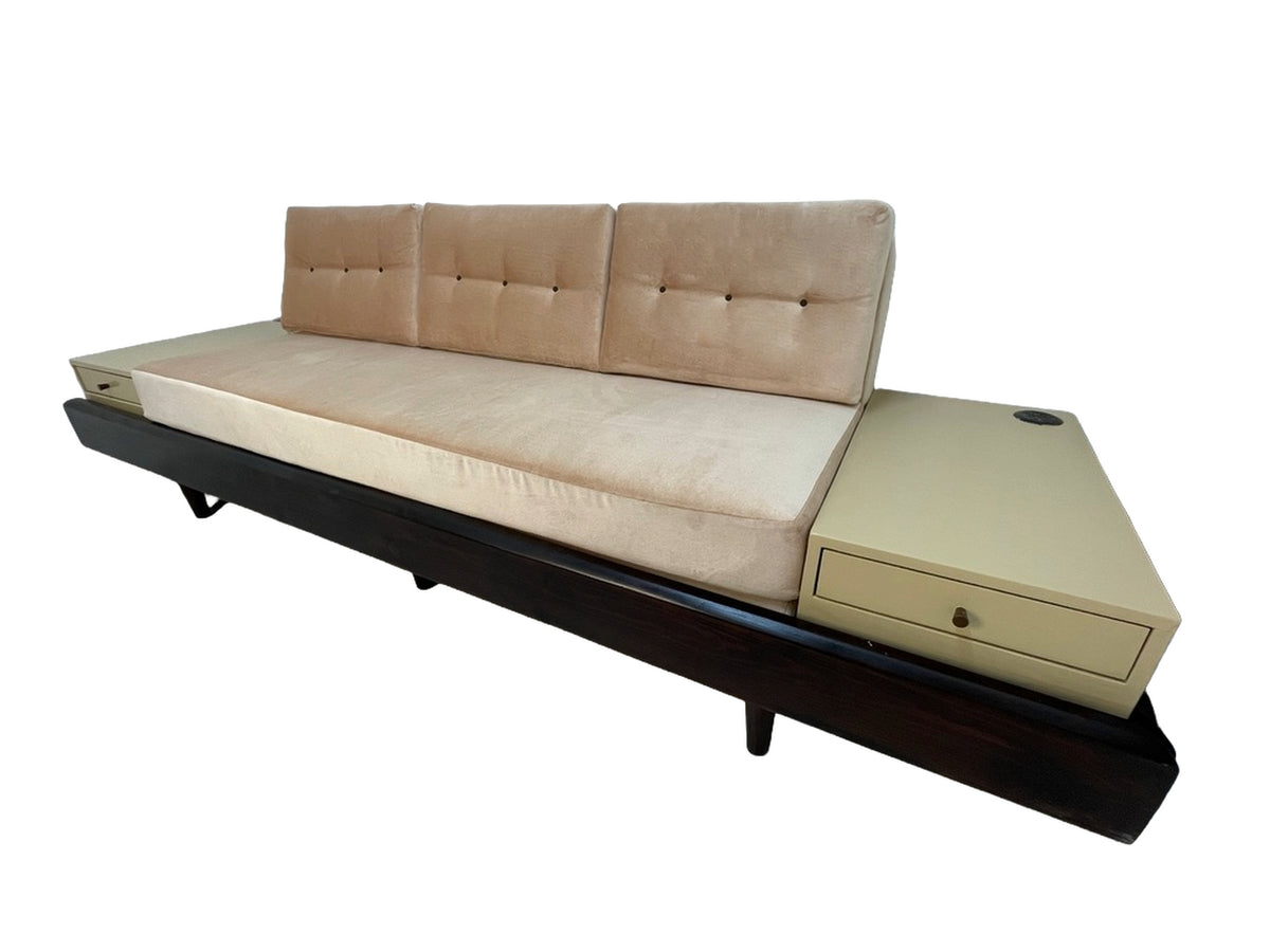 Mid-century style convertible sofa bed with blush upholstery, integrated side tables, storage drawers, and dark wood platform base viewed from the right.