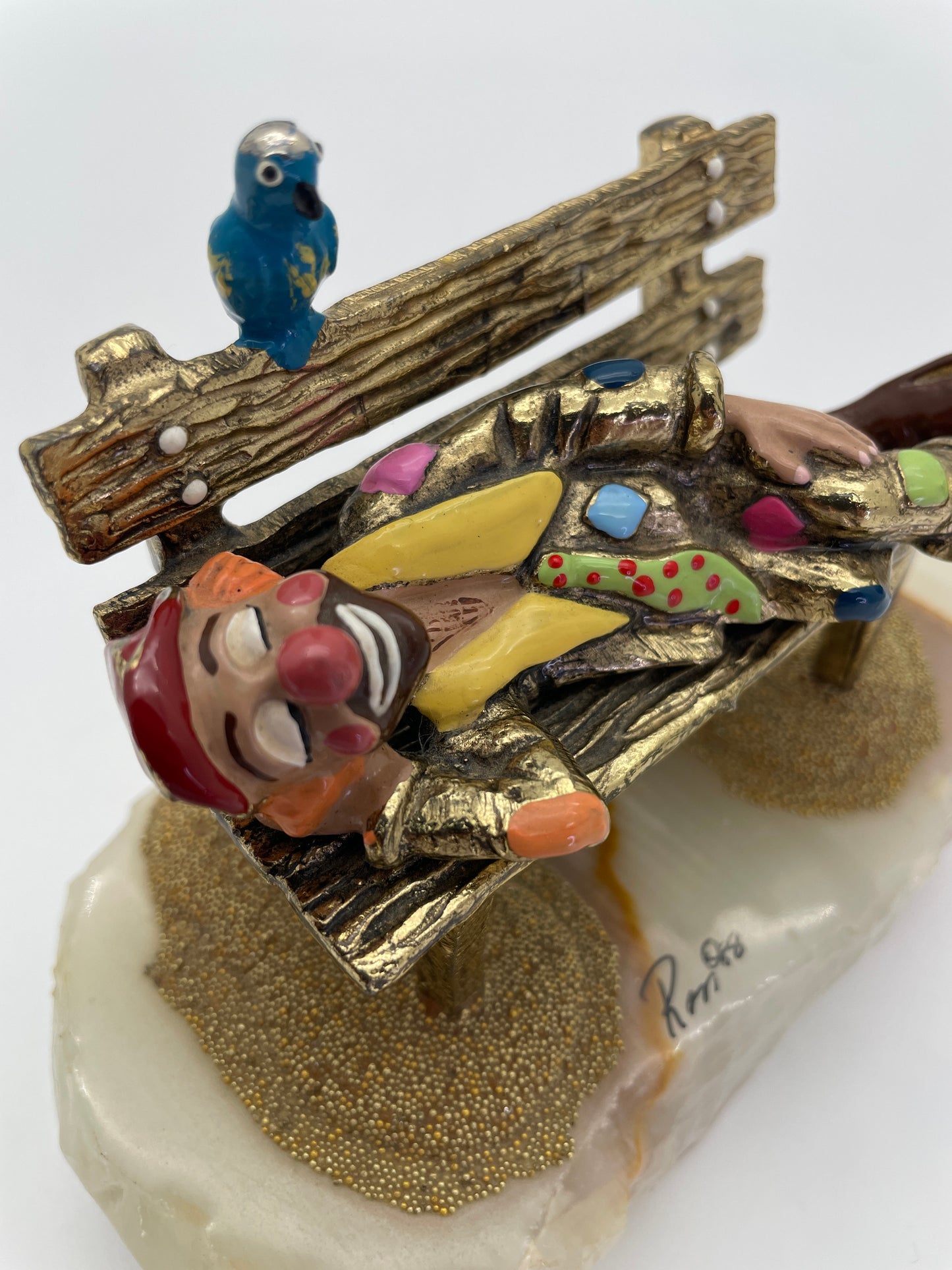 Detailed view of a Ron Lee sleeping clown sculpture highlighting the hand-painted costume, textured park bench, blue bird, natural onyx base, and Ron ’88 signature.