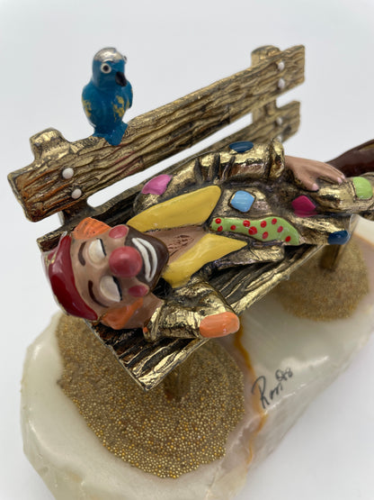 Detailed view of a Ron Lee sleeping clown sculpture highlighting the hand-painted costume, textured park bench, blue bird, natural onyx base, and Ron ’88 signature.
