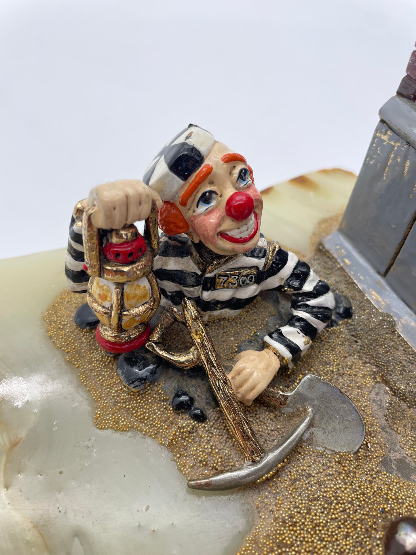 Close-up of the prisoner clown in Ron Lee’s Great Escape sculpture holding a lantern and pickaxe while emerging from a tunnel beneath a jail wall.