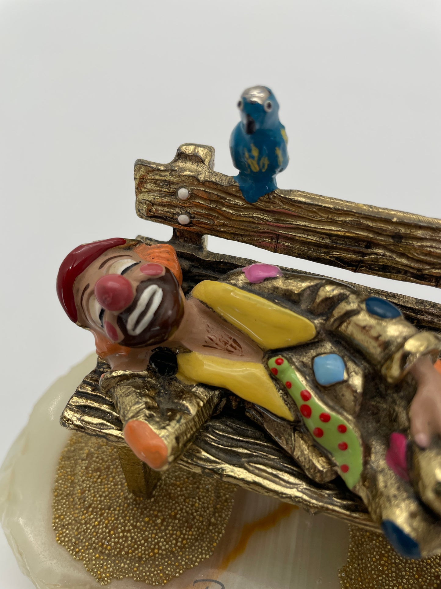 Close-up of a vintage Ron Lee sleeping clown sculpture showing the expressive hand-painted face, blue bird, rustic park bench, and signature 24K gold-plated finish.