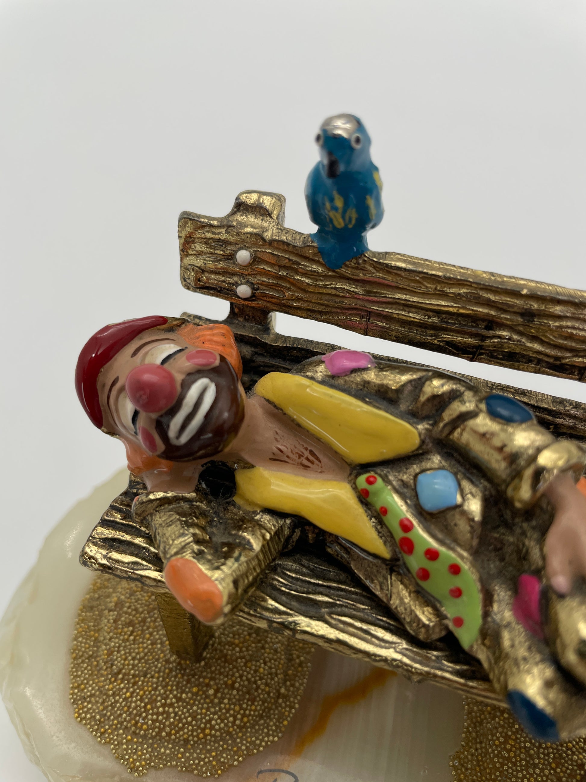Close-up of a vintage Ron Lee sleeping clown sculpture showing the expressive hand-painted face, blue bird, rustic park bench, and signature 24K gold-plated finish.