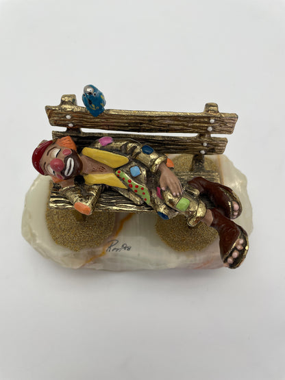 Overhead view of a vintage Ron Lee sleeping clown sculpture with blue bird, rustic park bench, hand-painted details, and natural onyx base.